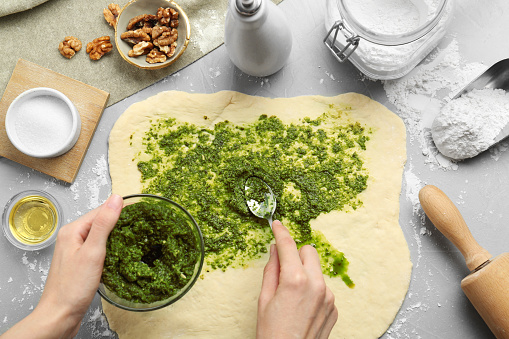 Making delicious pesto bread. Woman putting sauce onto raw dough at grey table, top view