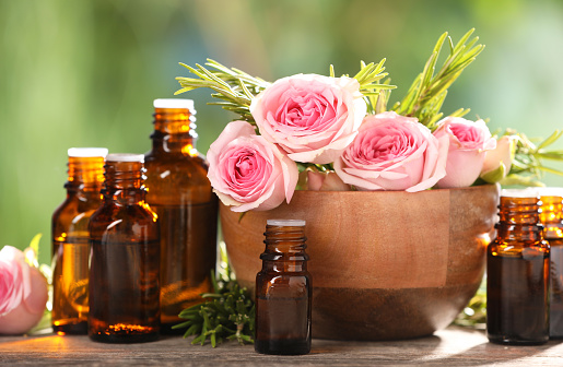 Bottles with essential oils, roses and rosemary on wooden table against blurred green background, closeup