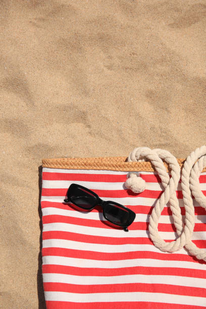 istock Striped beach bag and sunglasses on sand, top view. Space for text 1940627511