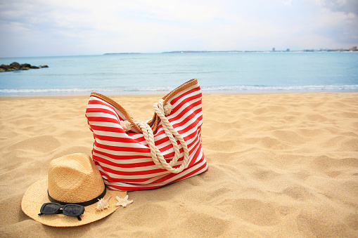 Stylish striped bag with straw hat, sunglasses, seashell and starfish on sandy beach near sea. Space for text