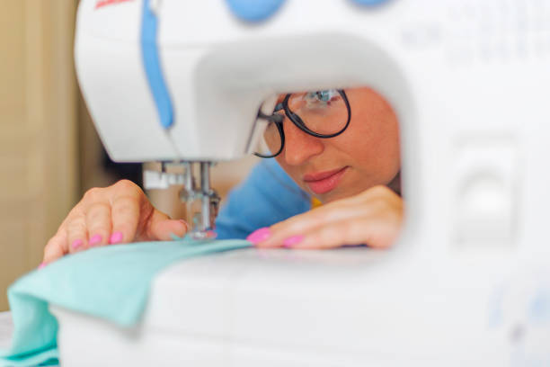 Focused woman with glasses sewing turquoise fabric on a white sewing machine, illustrating dedication to textile arts.