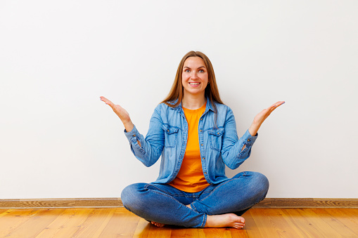 Smiling woman in blue jeans and denim jacket sitting cross-legged on the floor, shrugging with an amused expression.
