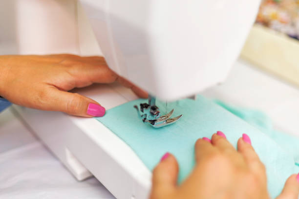 Back View Of A Senior Woman (80+) In Glasses Operating A Sewing Machine, Hands Guiding Blue Fabric On A Wooden Worktable Inside A Home Sewing Workshop, Illustrating Craftsmanship, Needlework Skills, DIY Creativity, And Active Aging.