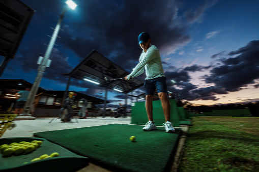 Low angle view looking up at a Pacific Islander male golf professional practicing his golf swing at the driving range on a cloudy evening in Hawaii.