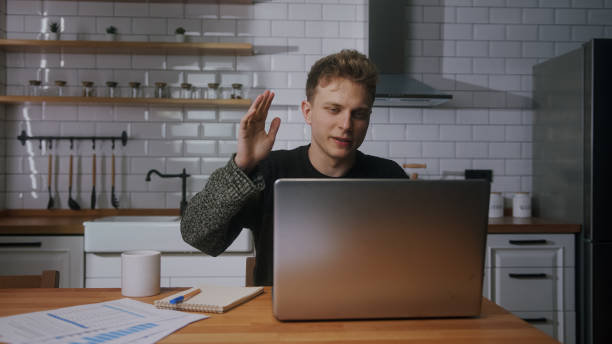 homme d’affaires blond assis à la maison dans la cuisine en train de regarder son ordinateur portable, de faire des vidéoconférences, de parler. travail à domicile, freelance, travail à distance, chat vidéo avec des collègues - colleague looking at camera indoors lifestyles photos et images de collection