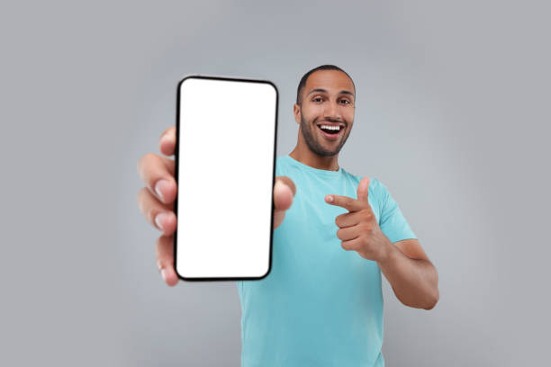 istock Young man showing smartphone in hand and pointing at it on light grey background 1940697961