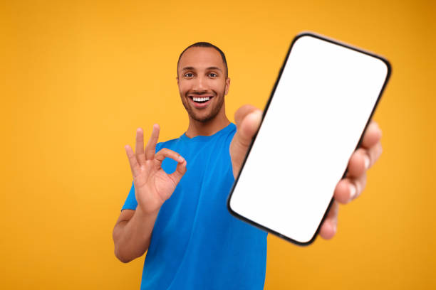 istock Young man showing smartphone in hand and OK gesture on yellow background 1940697977