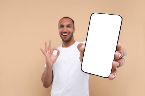 istock Young man showing smartphone in hand and OK gesture on beige background, selective focus. Mockup for design 1940699534