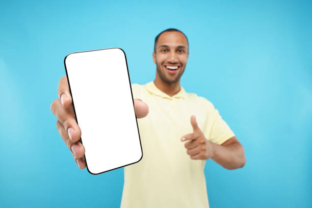 istock Young man showing smartphone in hand and pointing at it on light blue background, selective focus. Mockup for design 1940701095