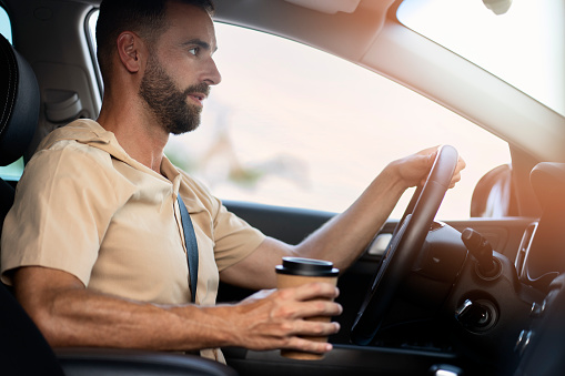 Handsome bearded latin man driving a new comfortable car holding cup of coffee. Transportation, road trip, car sharing concept