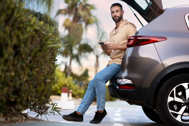 istock Latin man using mobile phone looking away planning road trip sitting on car trunk 1940716611