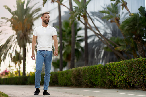 Confident handsome latin man wearing white t shirt, stylish blue jeans walking in park, looking away. Copy space