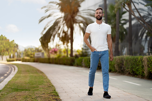 Smiling handsome man wearing white t shirt, stylish jeans walking on the street, looking away. Copy space. Vacation, travel concept