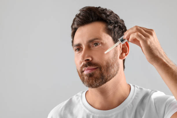 istock Handsome man applying cosmetic serum onto his face on light grey background. Space for text 1940744465