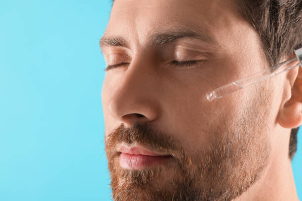 istock Handsome man applying cosmetic serum onto his face on light blue background, closeup 1940745504
