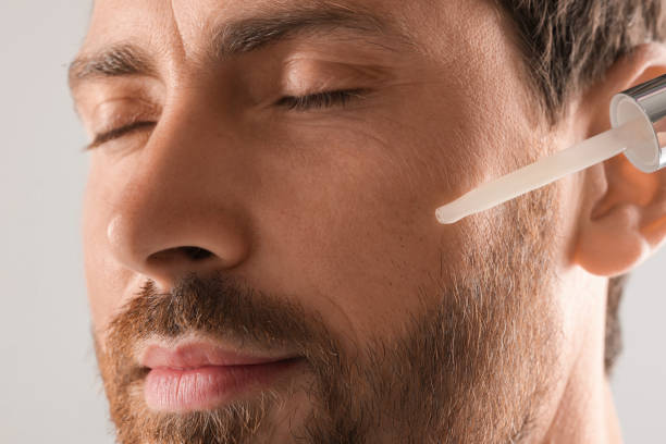 istock Handsome man applying cosmetic serum onto his face on light grey background, closeup 1940745505