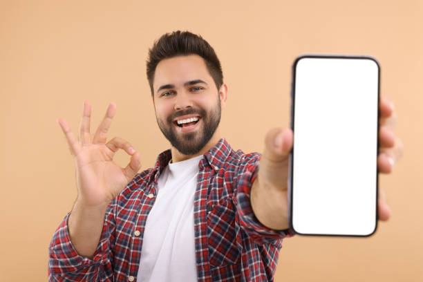 istock Young man showing smartphone in hand and OK gesture on beige background 1940762268