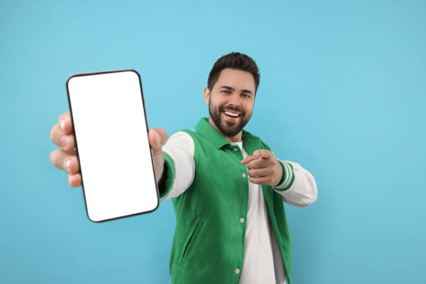 istock Young man showing smartphone in hand and pointing at camera on light blue background 1940762269