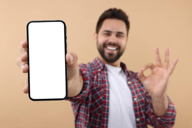 istock Young man showing smartphone in hand and OK gesture on beige background, selective focus. Mockup for design 1940764889