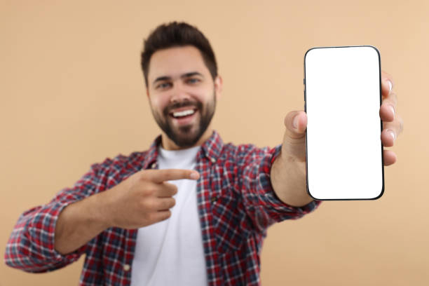 istock Young man showing smartphone in hand and pointing at it on beige background, selective focus. Mockup for design 1940765406