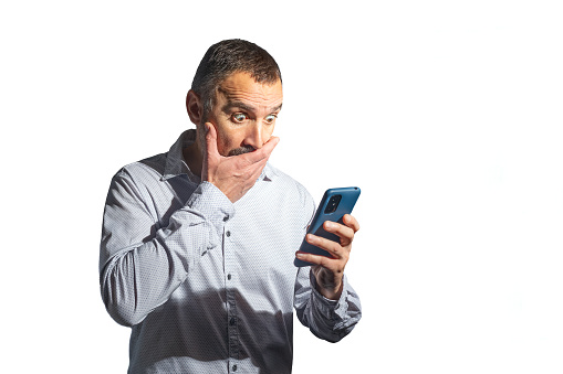 Madrid, Spain. Half-length view of a mature bearded man looking perplexed and incredulous at the screen of his mobile phone. Isolated against a white background.