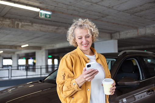 A smiling mid-adult woman is standing by her car in a parking spot, drinking coffee and using a smart phone