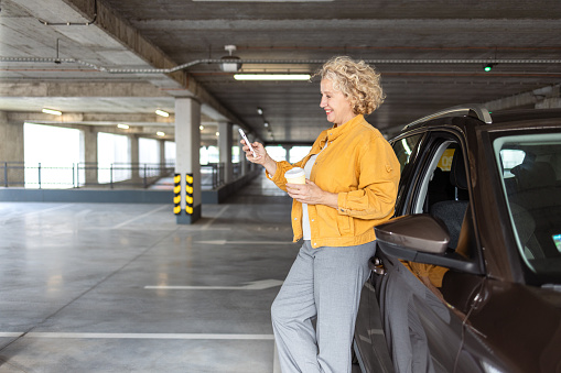 A mid-adult woman using a smart phone while leaning on the car. She parked a car to have a coffee break.