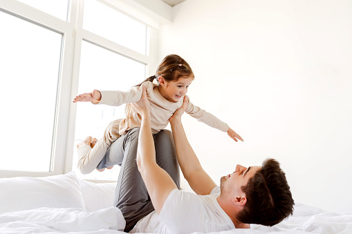 Portrait of smiling father holding little happy daughter fooling around lying on bed together in cozy room at home. Positive family. Childhood concept