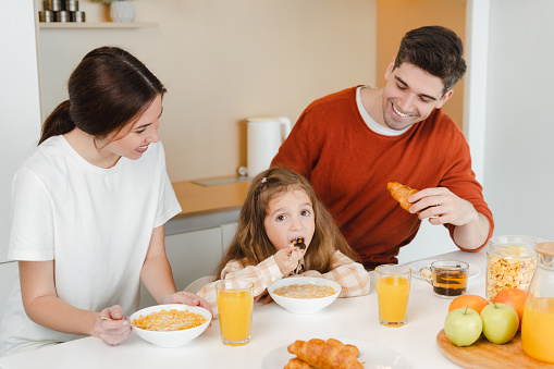 Portrait of happy, smiling family, mother, father and little daughter eating together in kitchen at home. Positive family together in morning sitting at table. Food concept