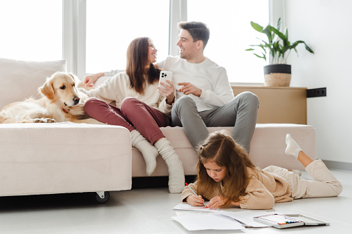 Portrait of smiling happy mother and father, woman holding mobile phone, dog lying on sofa near them, little daughter sitting on floor drawing, in cozy home. Concept of parenthood, online shopping