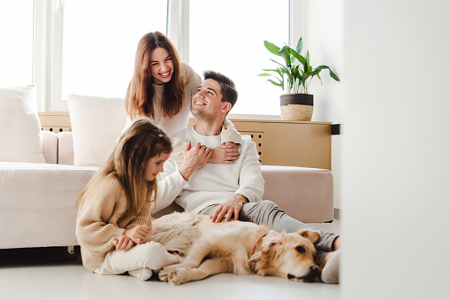 Smiling beautiful family, young mother, father and little daughter relaxing together, woman hugging man, little girl petting and playing with dog, golden retriever. Concept of love, parenthood