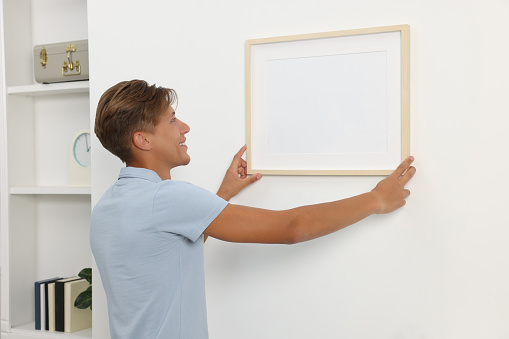 Young man hanging picture frame on white wall indoors