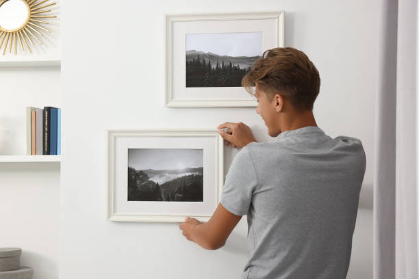 istock Young man hanging picture frames on white wall indoors, back view 1940792659