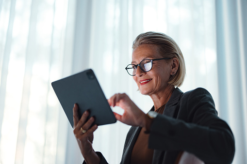 Close up shot of a beautiful successful senior woman in business wearing glasses. She is standing, holding a digital tablet, presenting or leading a meeting while smiling and looking down.