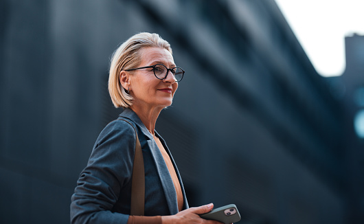 Portrait of a beautiful senior business woman wearing glasses and formal clothes, on her way to the meeting or to the office. She is walking on the streets, holding a mobile phone, looking away and smiling.
