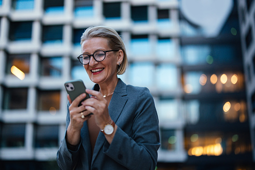 Close up shot of an older business woman standing in front of an office building, texting on her phone in the evening. She is looking down and smiling.