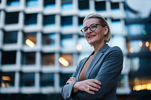 Close up shot of a distinguished older business woman wearing formal clothes and glasses. She is standing in front of the office building with her arms crossed, smiling and looking away.
