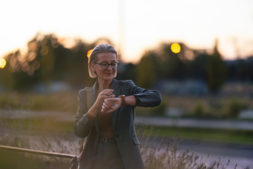 Sunset portrait of a beautiful senior business woman standing by the fence checking her wrist watch while waiting for someone to meet.