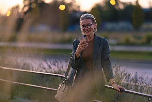 Portrait of a cheerful older business woman wearing glasses, standing outdoors in a beautiful evening light, waiting for someone, texting on the mobile phone and smiling.