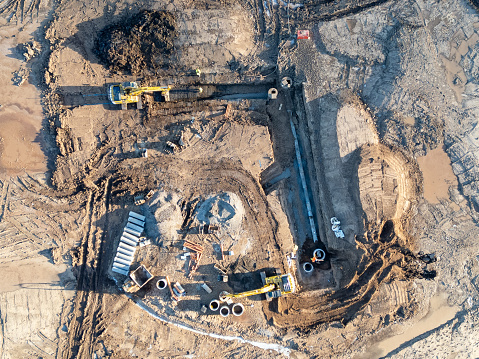 Aerial drone view of a housing development construction site in Scotland.  Two bulldozers are at work, digging trenches for concrete pipes and drains, in preparation for the building of new houses. The heavy machinery is seen directly below.