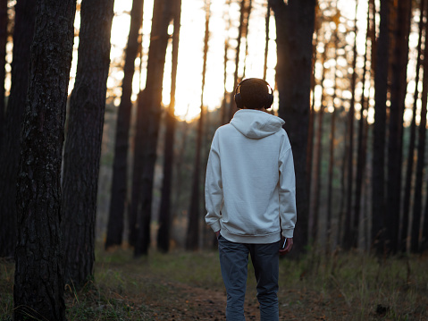 A young man in a white hoodie walks through the forest at sunset and listens to music with headphones. The concept of freedom. Alone man