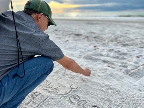 Active senior man squats down to write a message in the white sand at Indian Rocks beach, Florida on New Years Day against sand and sky near the time of sunset.