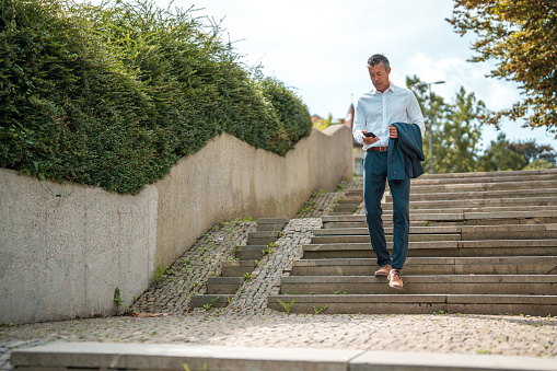 Asian businessman in his mid-40s navigating the stairs of a public park while engaged with his smartphone.