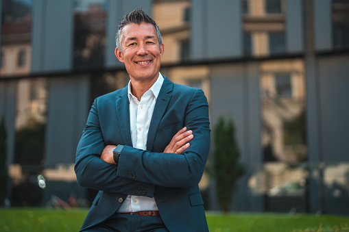 A mid adult Asian businessman standing with arms crossed beside a corporate building in his sleek suit.