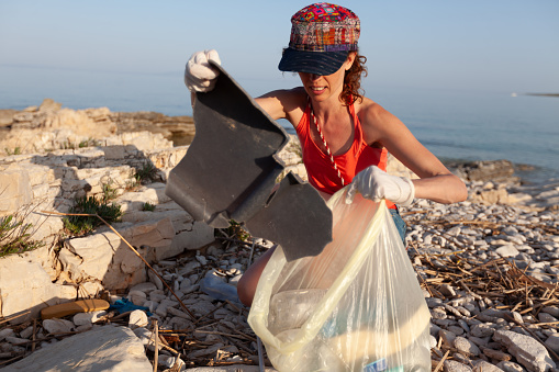 Front View of Young Adult Caucasian Woman in Protective Gloves Collecting Garbage in Squatting Position on a Wild Beach Postponing an Environment Disaster
