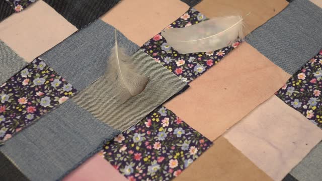 A swan feather falls on a colorful vintage textile patchwork background with denim elements in blue tones, slow motion