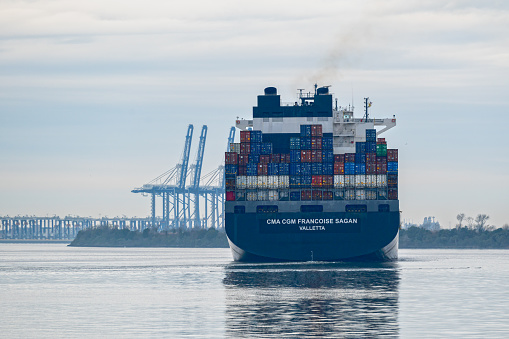 Charleston, SC, USA - January 16, 2024: Stern of the 304-meter container ship CMA CGM Francoise Sagan sailing into Charleston Harbor with ship-to-shore cranes of Leatherman Terminal in the background.