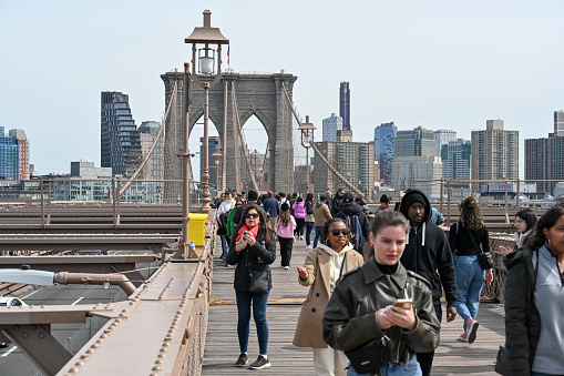 New York City, New York, USA, April 8, 2023 - Tourists and locals walk across the Brooklyn Bridge, with the Brooklyn skyline in the background.