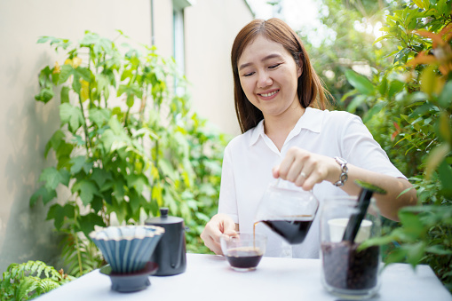 Happy Asian beautiful woman making a specialty coffee in morning at her backyard garden, woman brewing a coffee by dripping or pouring over a hot water.