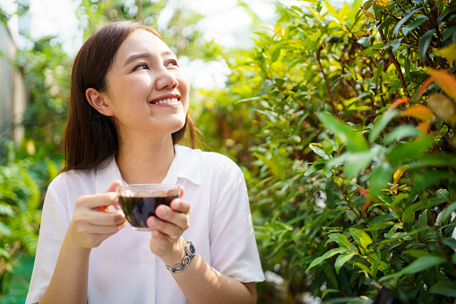Asian beautiful woman enjoy drinking a cup of coffee in morning at home backyard. Smiling cheerful woman drinks a black coffee in the nature close up.